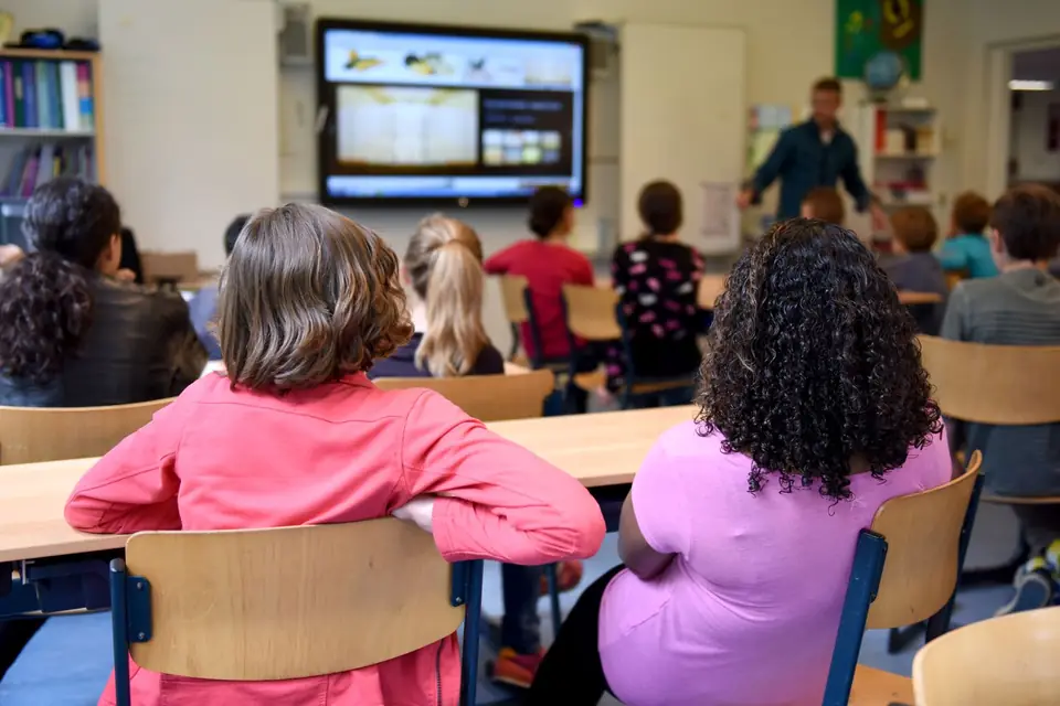 Leerkracht geeft les bij het digibord terwijl de leerlingen van een basisschoolklas meekijken