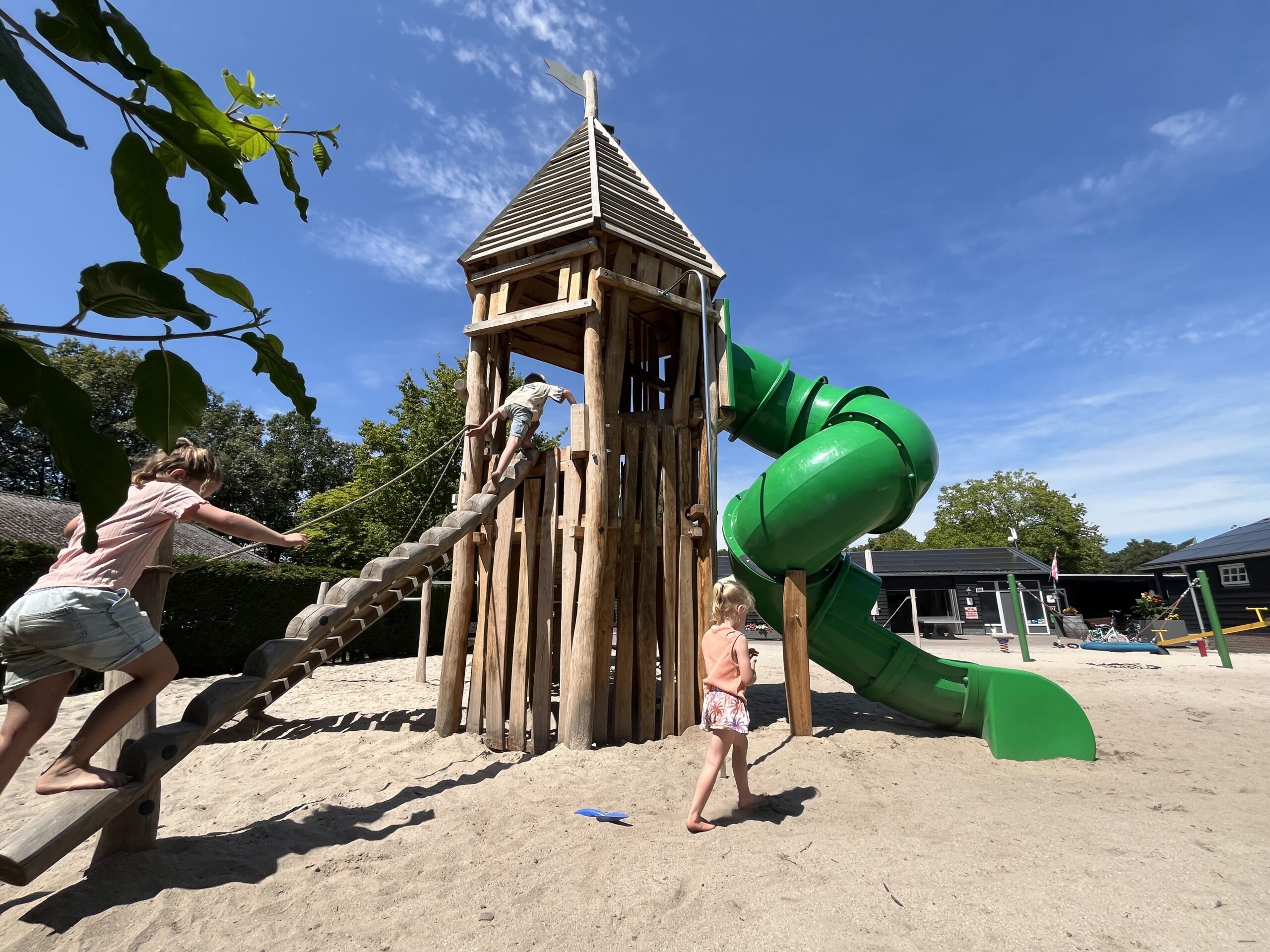 Drie kinderen spelen op de zandige speeltuin met houten toren en groene spiraalglijbaan op Camping De Peel bij Asten