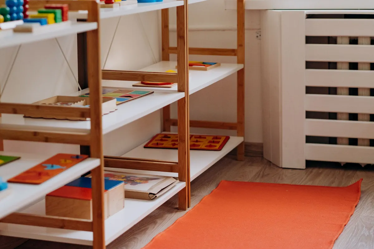 Houten rekken met educatief speelgoed en een oranje speelmat in een lichte kinderopvangruimte